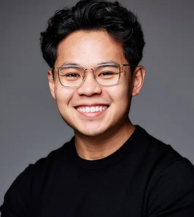 Young man wearing glasses and black shirt smiling at camera against gray background.