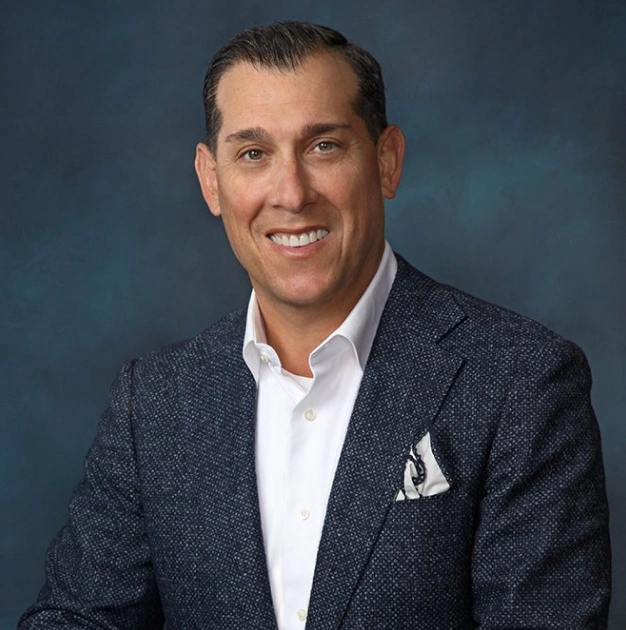 Professional headshot of man in dark blazer and white shirt against blue background for dental practice.