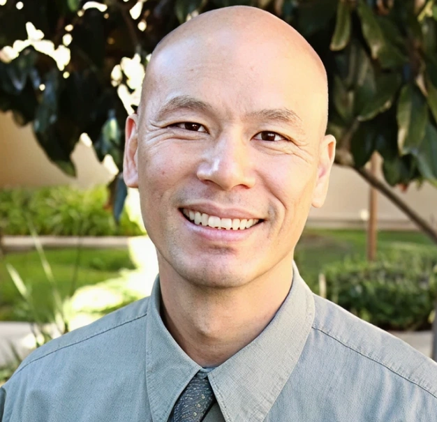 Professional headshot of smiling man in gray shirt and tie against outdoor garden background.