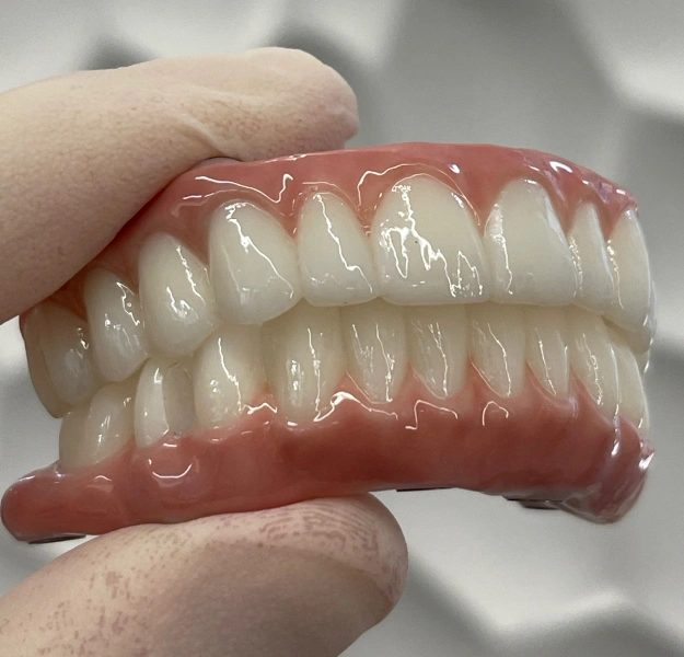 Hand holding complete denture with full set of artificial teeth against blurred background.